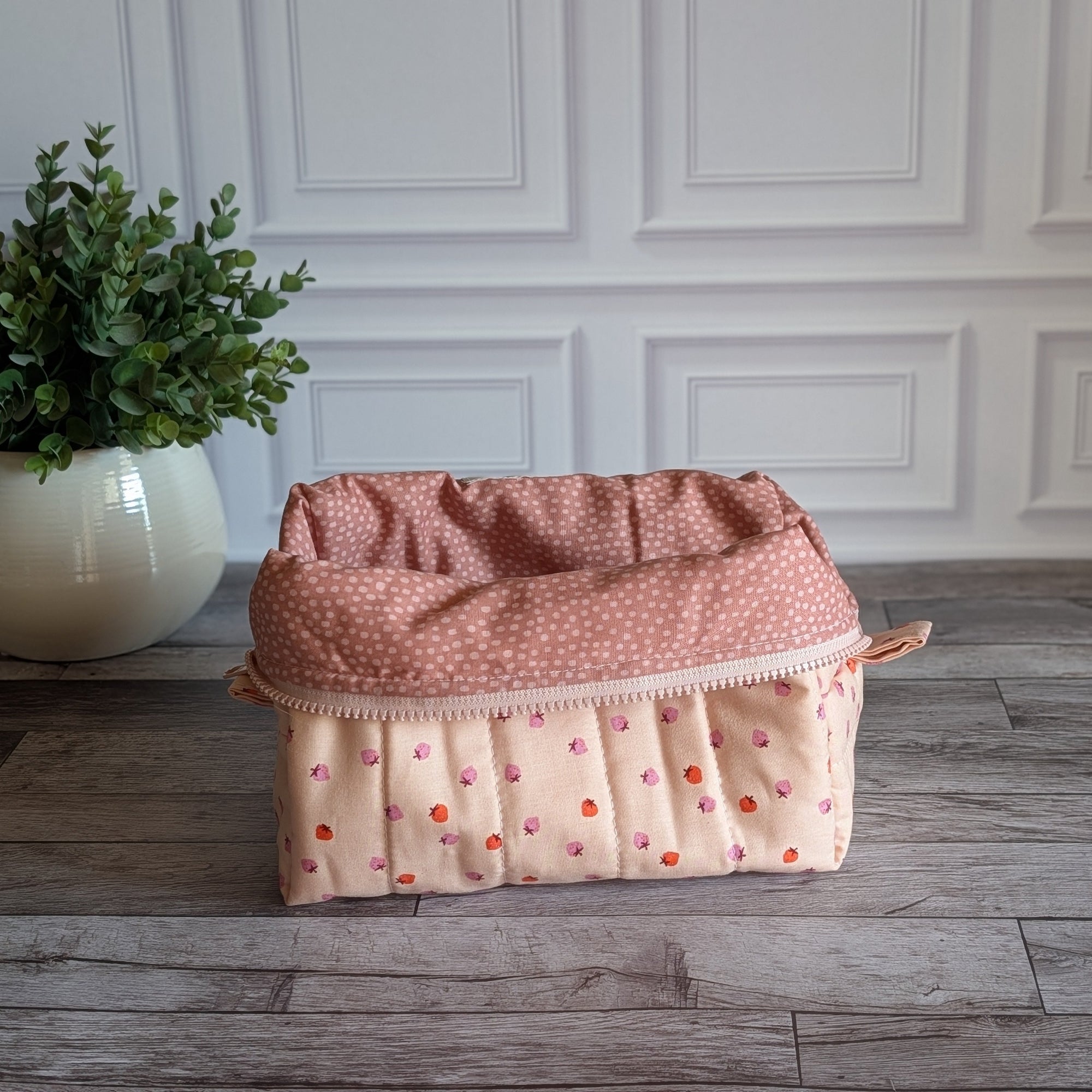 A quilted cosmetic pouch in a light peach-pink color with a pattern of small orange and pink strawberries, standing on a gray wood floor next to a potted green plant.