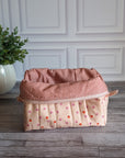 A quilted cosmetic pouch in a light peach-pink color with a pattern of small orange and pink strawberries, standing on a gray wood floor next to a potted green plant.