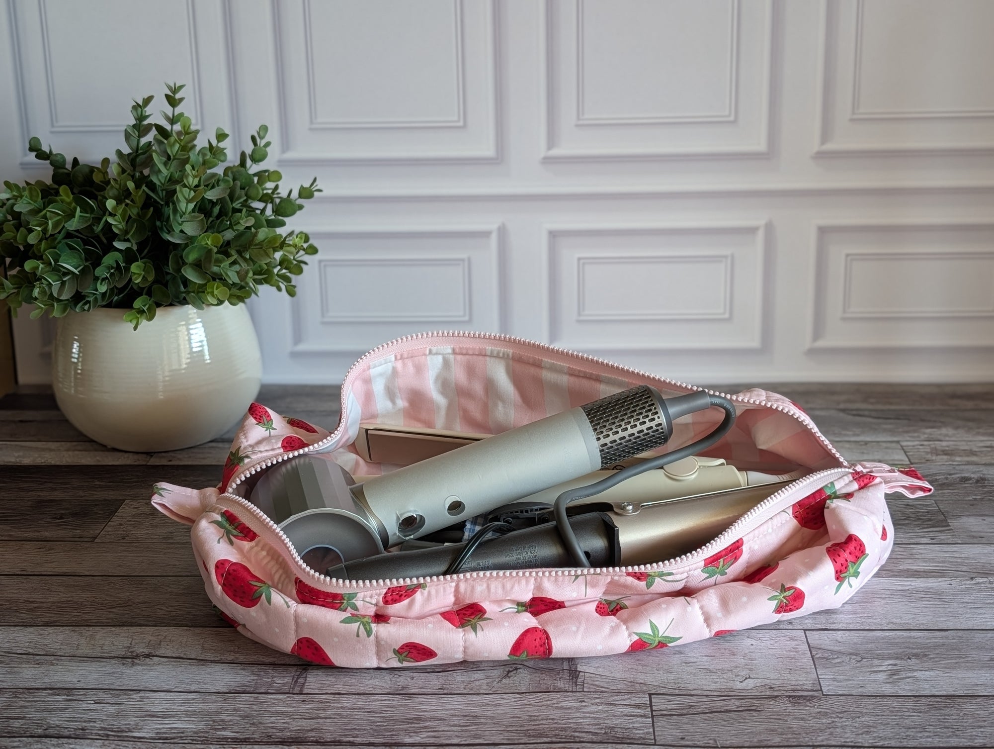 A pink quilted cosmetic pouch with a strawberry print is open on a gray wood floor, revealing hair styling tools inside. A potted green plant is visible in the background against a white paneled wall.