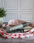 A pink quilted cosmetic pouch with a strawberry print is open on a gray wood floor, revealing hair styling tools inside. A potted green plant is visible in the background against a white paneled wall.
