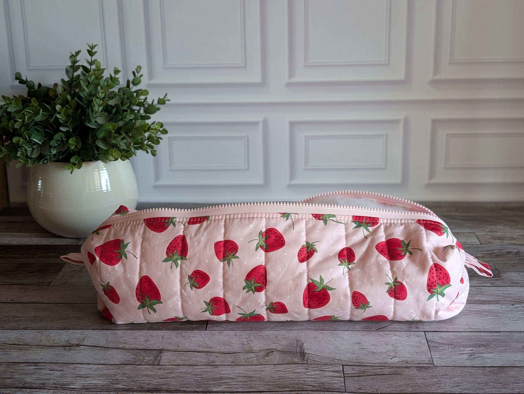 A quilted cosmetic pouch with a pink and red strawberry pattern, resting on a gray wooden floor in front of a white paneled wall with a potted green plant in the background.