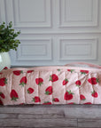 A quilted cosmetic pouch with a pink and red strawberry pattern, resting on a gray wooden floor in front of a white paneled wall with a potted green plant in the background.