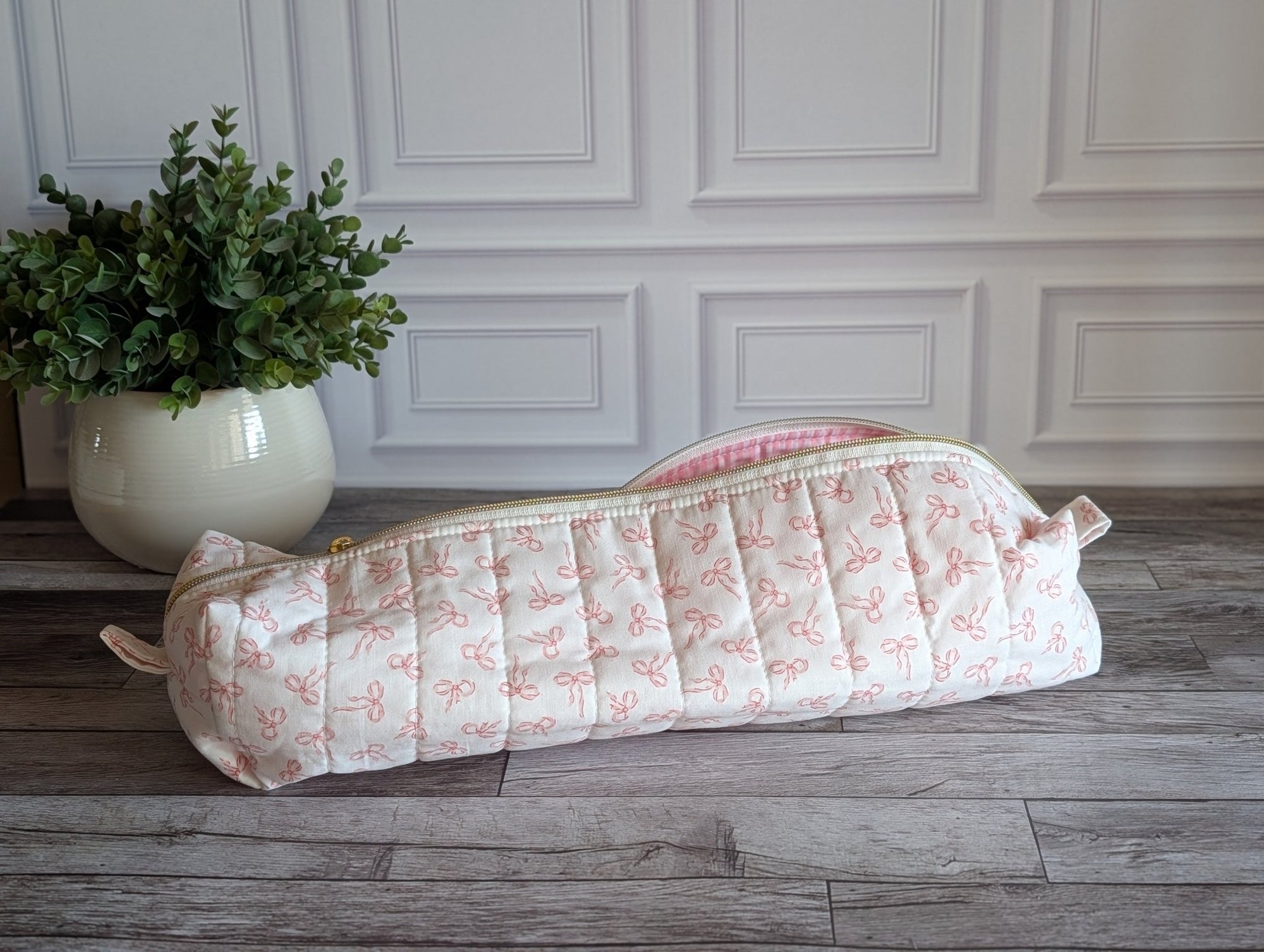 A quilted fabric zippered pouch with a pink bow pattern on a cream background, resting on a gray wood floor in front of a white paneled wall with a potted green plant.