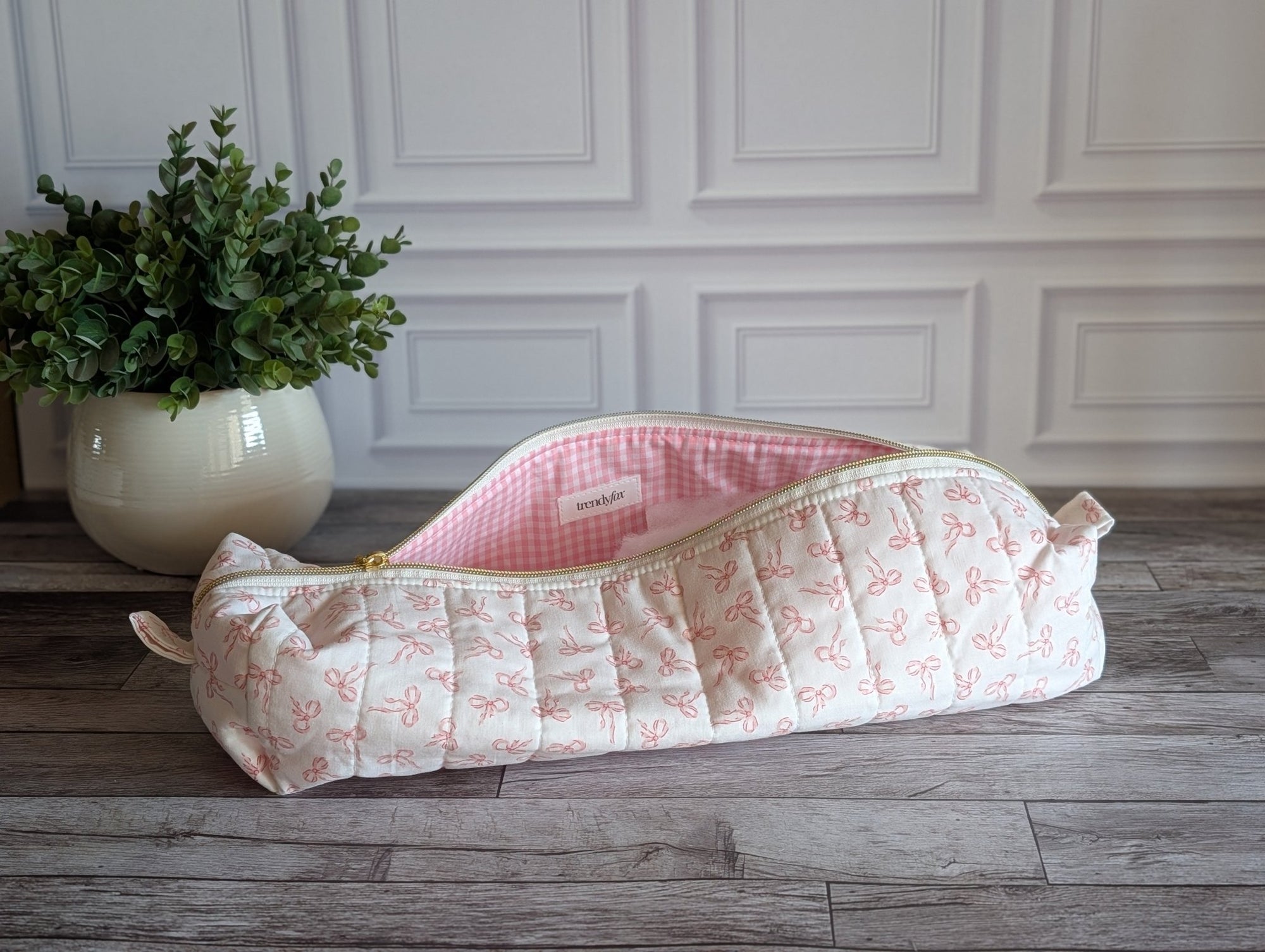 A quilted cosmetic pouch with a pink and white floral print and a pink gingham lining, resting on a gray wooden floor in front of a white paneled wall.