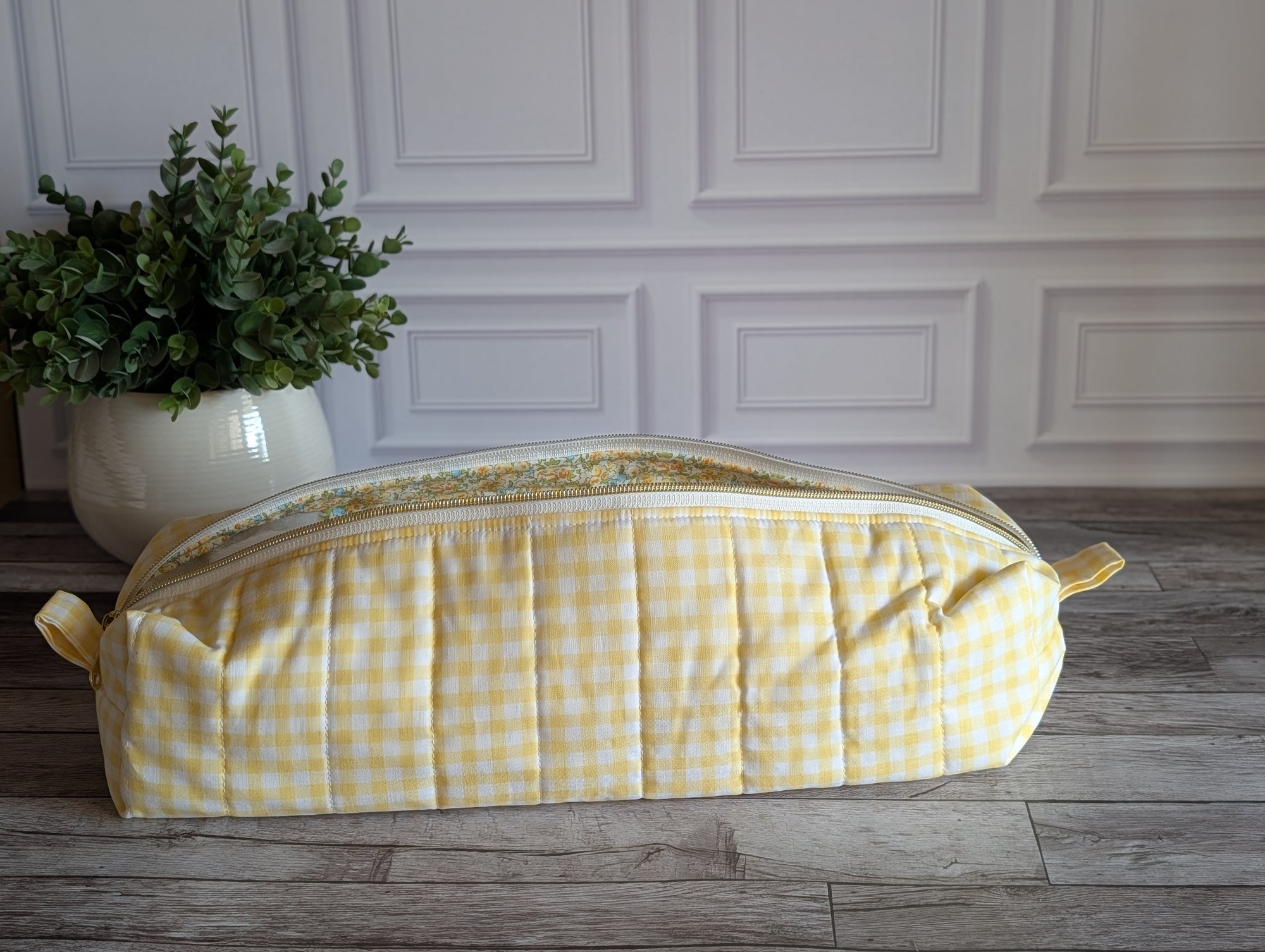A yellow and white gingham quilted cosmetic bag with a floral patterned interior lining, resting on a gray wooden floor in front of a white paneled wall.