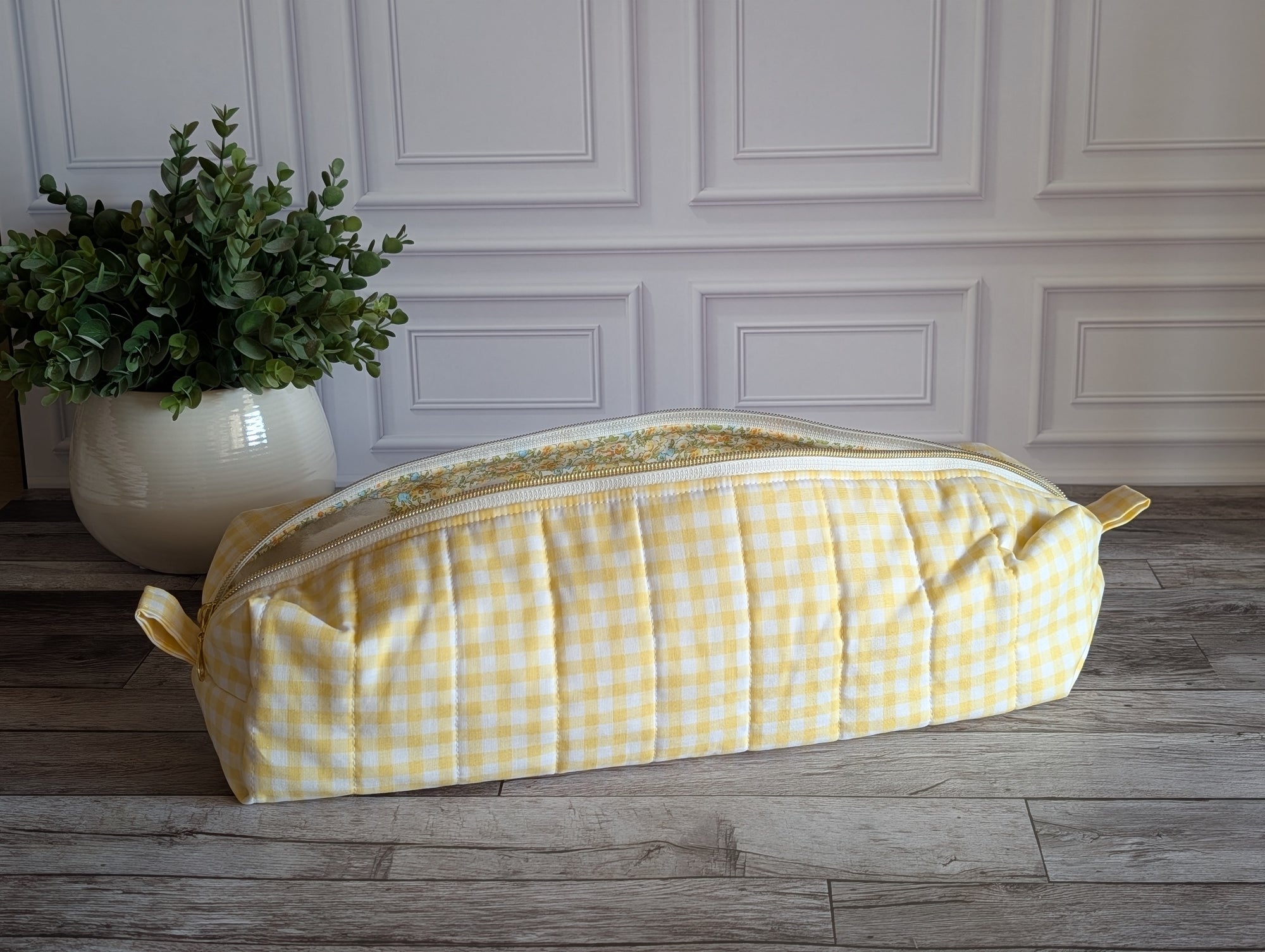 A yellow and white gingham quilted cosmetic bag with a floral patterned lining, resting on a gray wooden floor in front of a white paneled wall.