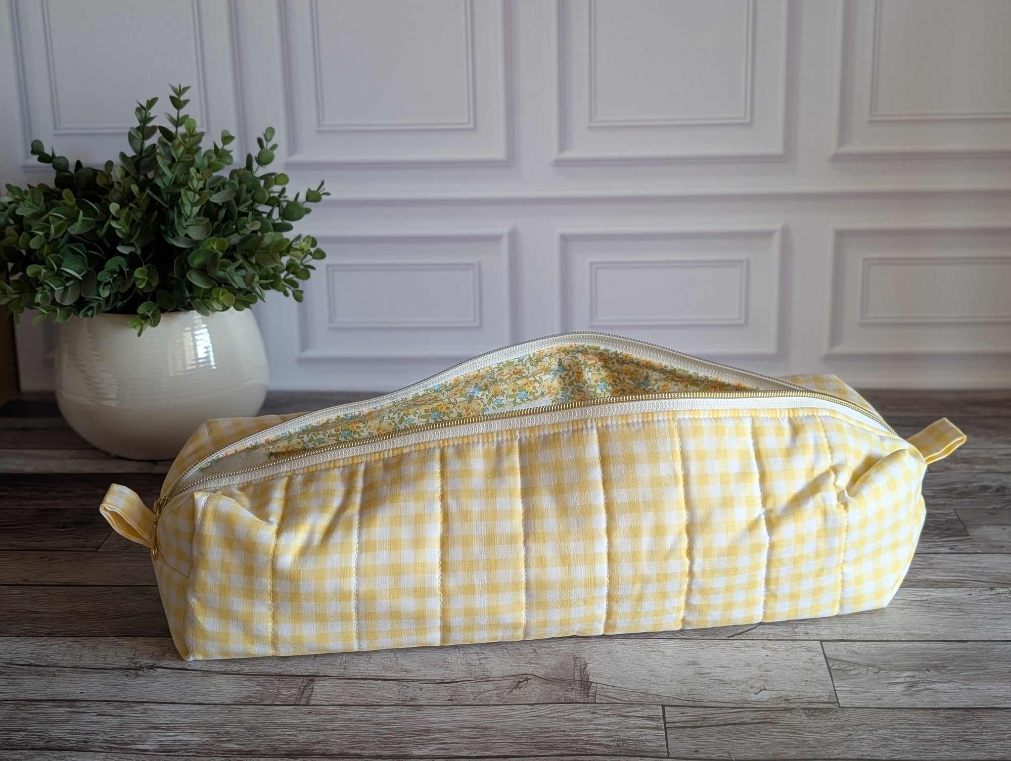 A yellow and white gingham quilted travel or toiletry bag with a floral patterned lining, resting on a rustic wooden floor in front of a white paneled wall.