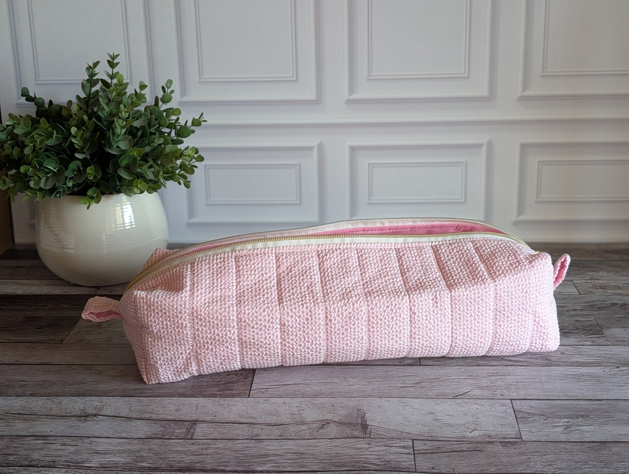 A light pink quilted fabric zippered pouch or cosmetic bag resting on a gray wood floor in front of a white paneled wall, with a potted green plant on the left.