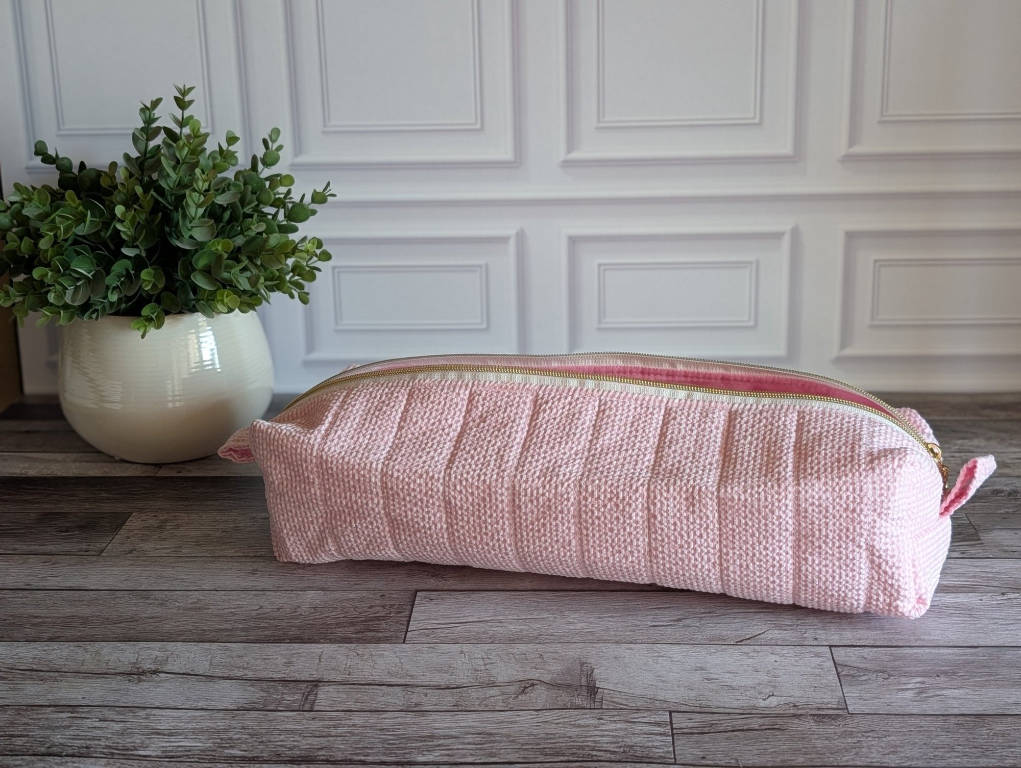 A light pink textured zippered pouch or cosmetic bag resting on a rustic wooden floor in front of a white paneled wall, with a potted green plant on the left.
