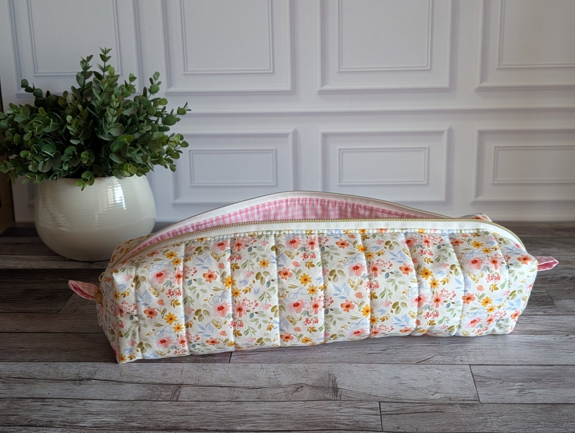 A quilted fabric cosmetic pouch with a floral print and pink gingham trim, resting on a gray wooden floor in front of a white paneled wall with a potted plant in the background.