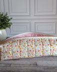 A quilted fabric cosmetic pouch with a floral print and pink gingham trim, resting on a gray wooden floor in front of a white paneled wall with a potted plant in the background.