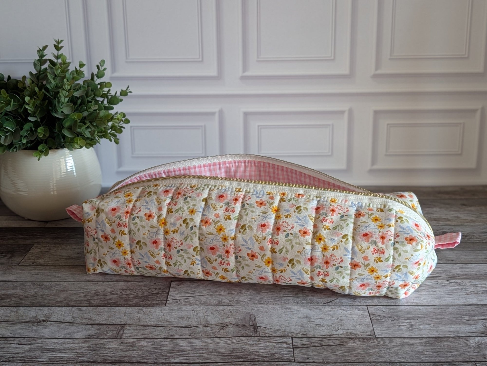 A quilted cosmetic pouch with a floral print and pink gingham trim, resting on a gray wooden floor in front of a white paneled wall with a potted plant to the side.