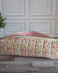 A quilted cosmetic pouch with a floral print and pink gingham trim, resting on a gray wooden floor in front of a white paneled wall with a potted plant to the side.
