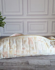 A quilted fabric pencil case with a pastel floral and bow pattern, resting on a gray wood floor in front of a white paneled wall with a potted plant on the left.