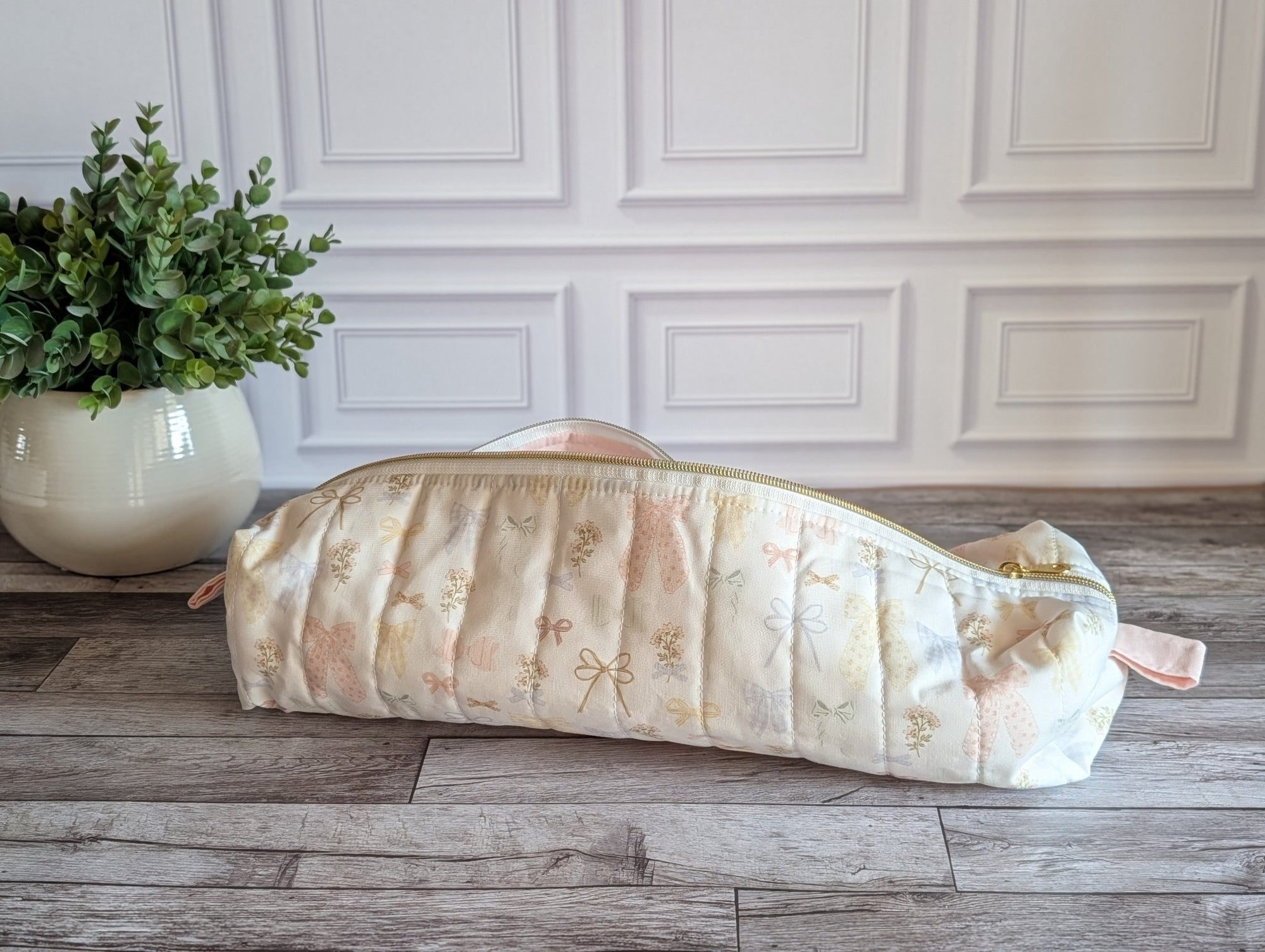A quilted pencil case with a cream and pale pink floral pattern, resting on a gray wood floor in front of a white paneled wall.