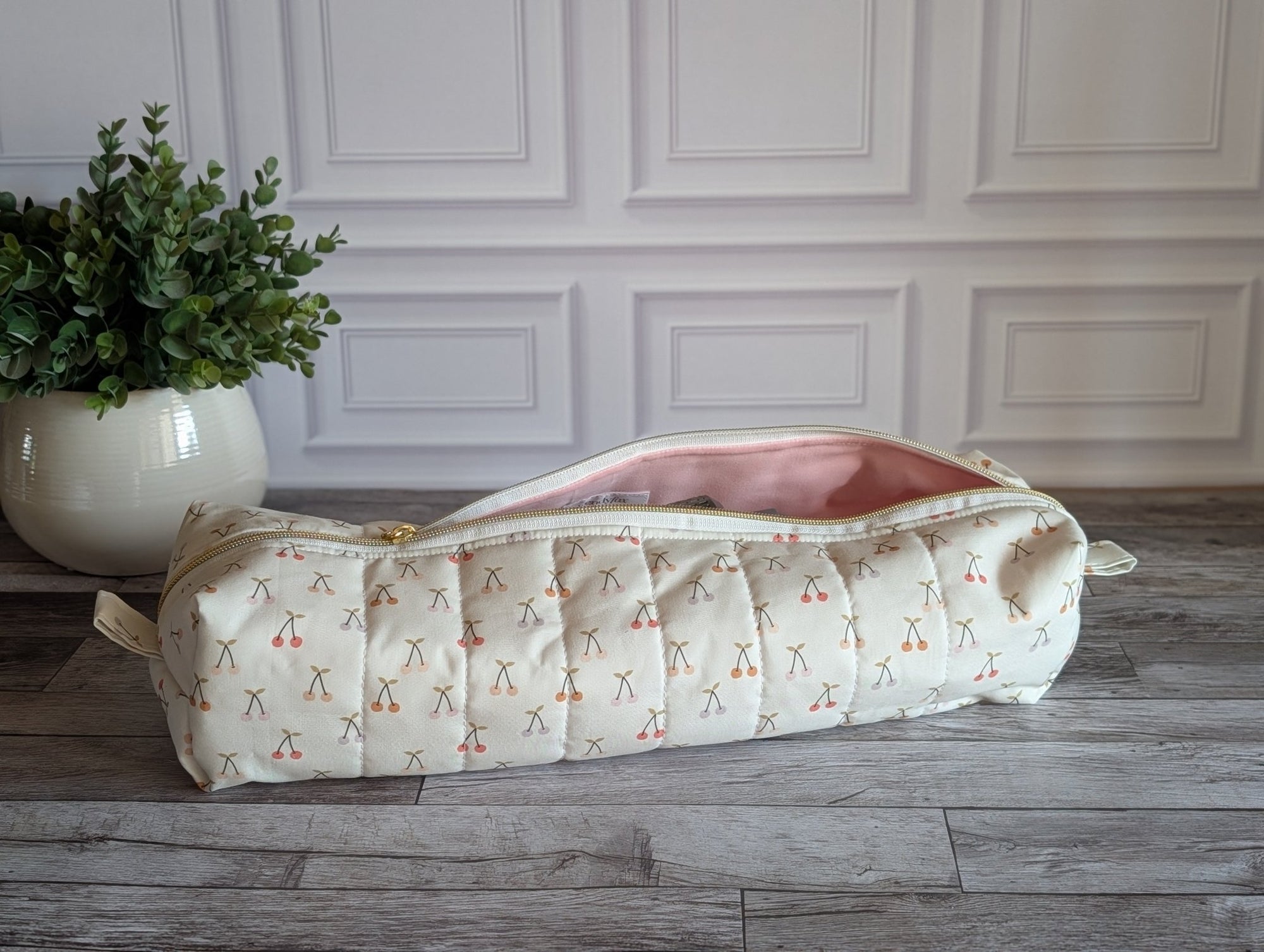 A quilted cylindrical cosmetic pouch with a cream and pink cherry pattern, shown open on a gray wood floor in front of a white paneled wall.