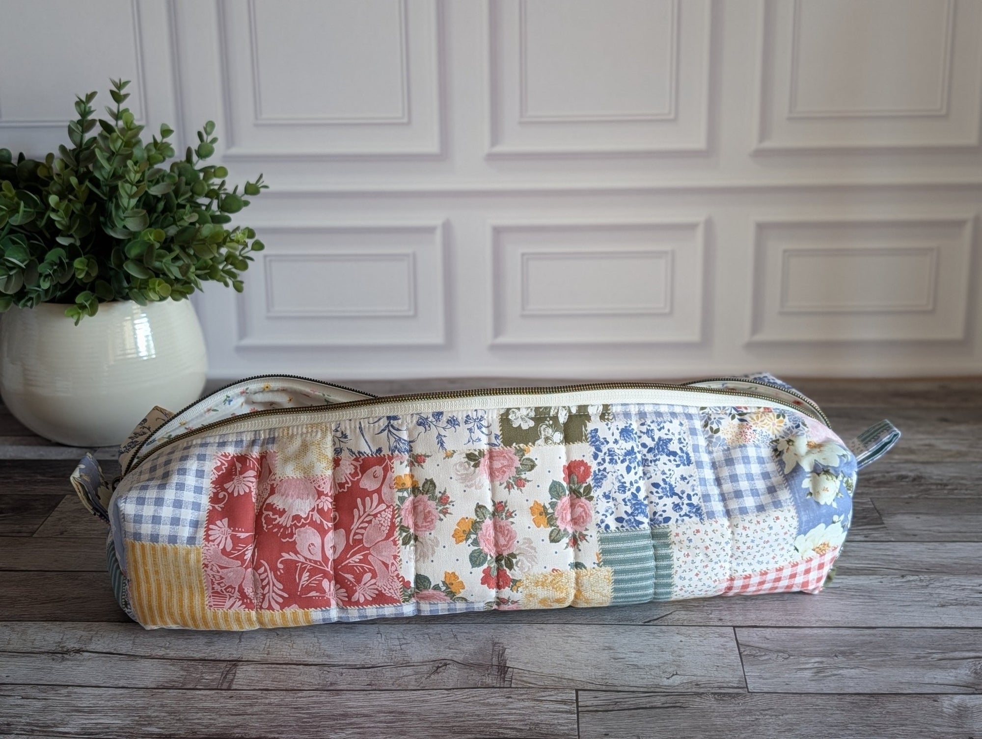 A long rectangular quilted patchwork zippered pouch or case with a floral and gingham pattern, resting on a gray wood floor in front of a white paneled wall with a potted green plant on the left.