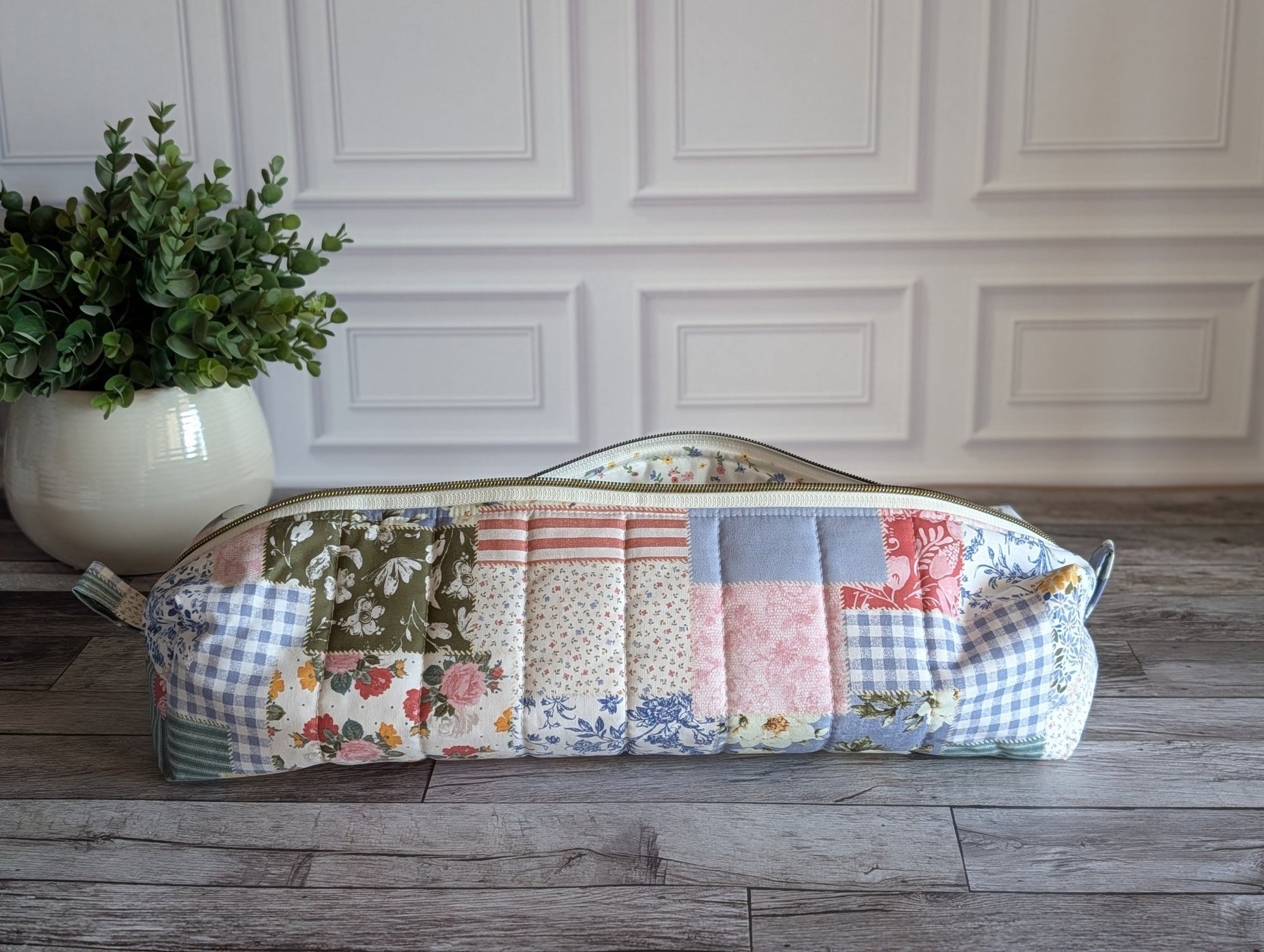 A long quilted patchwork zipper pouch or case with floral and gingham fabric panels, resting on a gray wood floor in front of a white paneled wall with a potted green plant on the left.