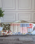 A long quilted patchwork zipper pouch or case with floral and gingham fabric panels, resting on a gray wood floor in front of a white paneled wall with a potted green plant on the left.