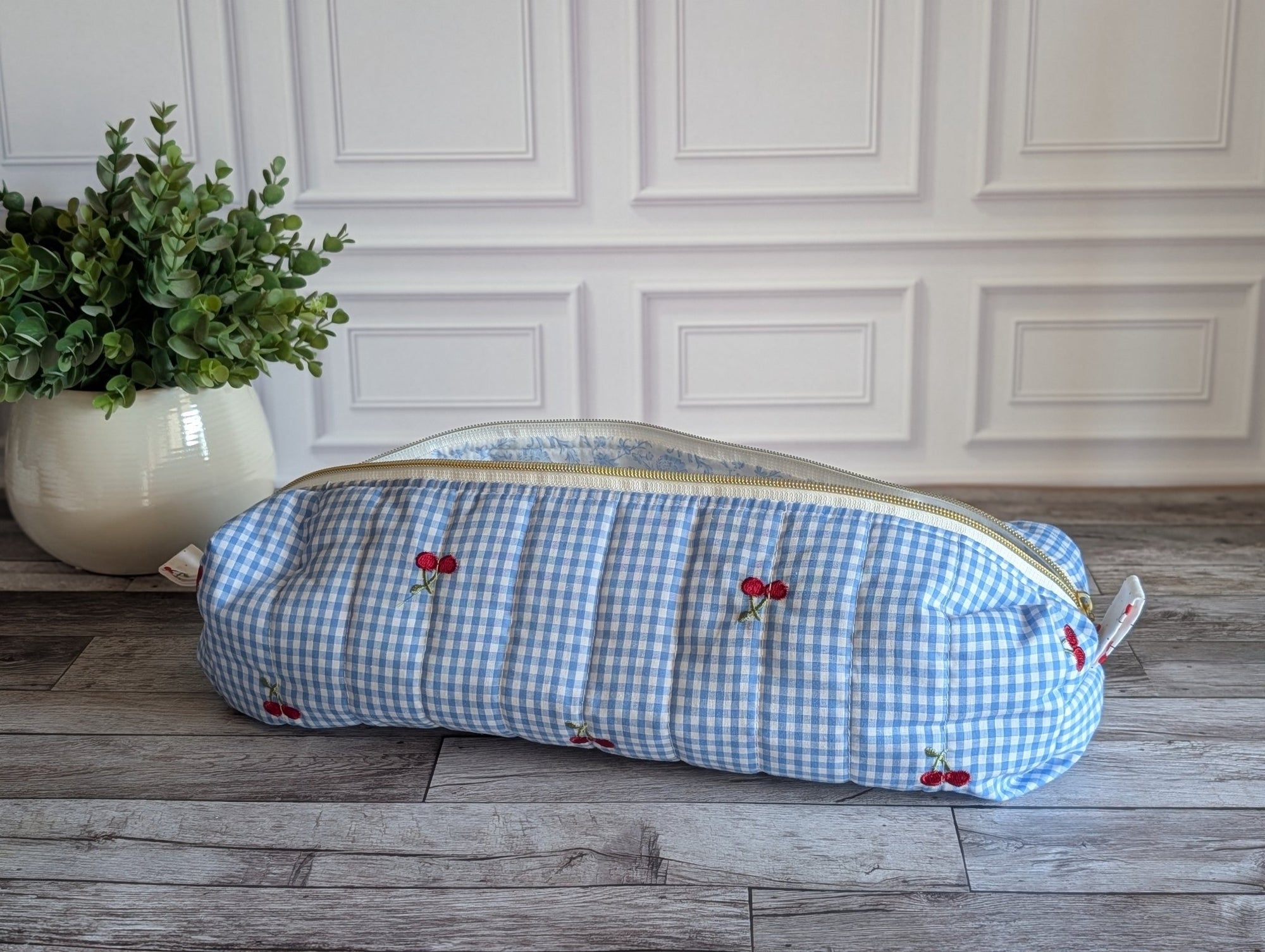 A light blue and white gingham zippered pouch with embroidered red cherries, resting on a gray wood floor next to a potted green plant.