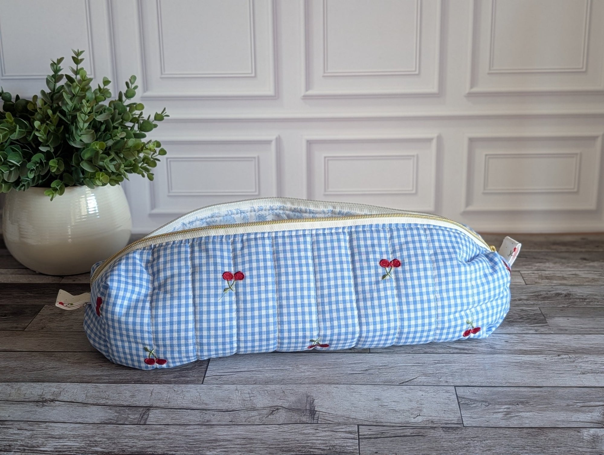 A light blue and white gingham zippered pouch with small red cherry prints, resting on a gray wooden floor in front of a white paneled wall.
