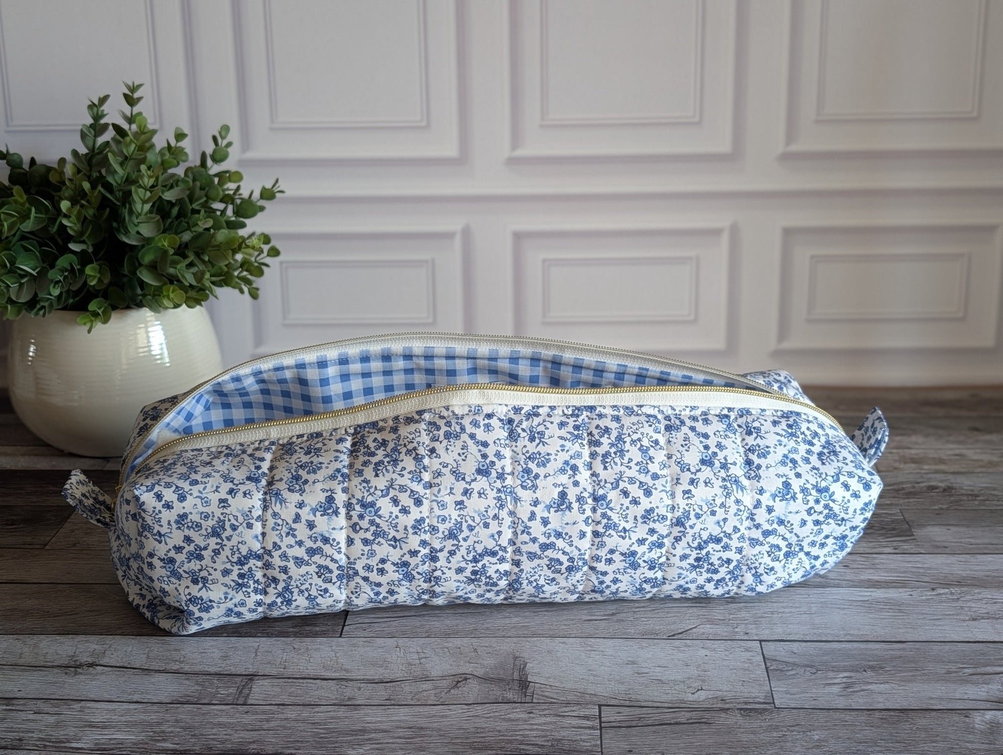 A quilted fabric storage bag with a blue and white floral print exterior and a blue and white gingham lining, resting on a gray wood floor in front of a white paneled wall.