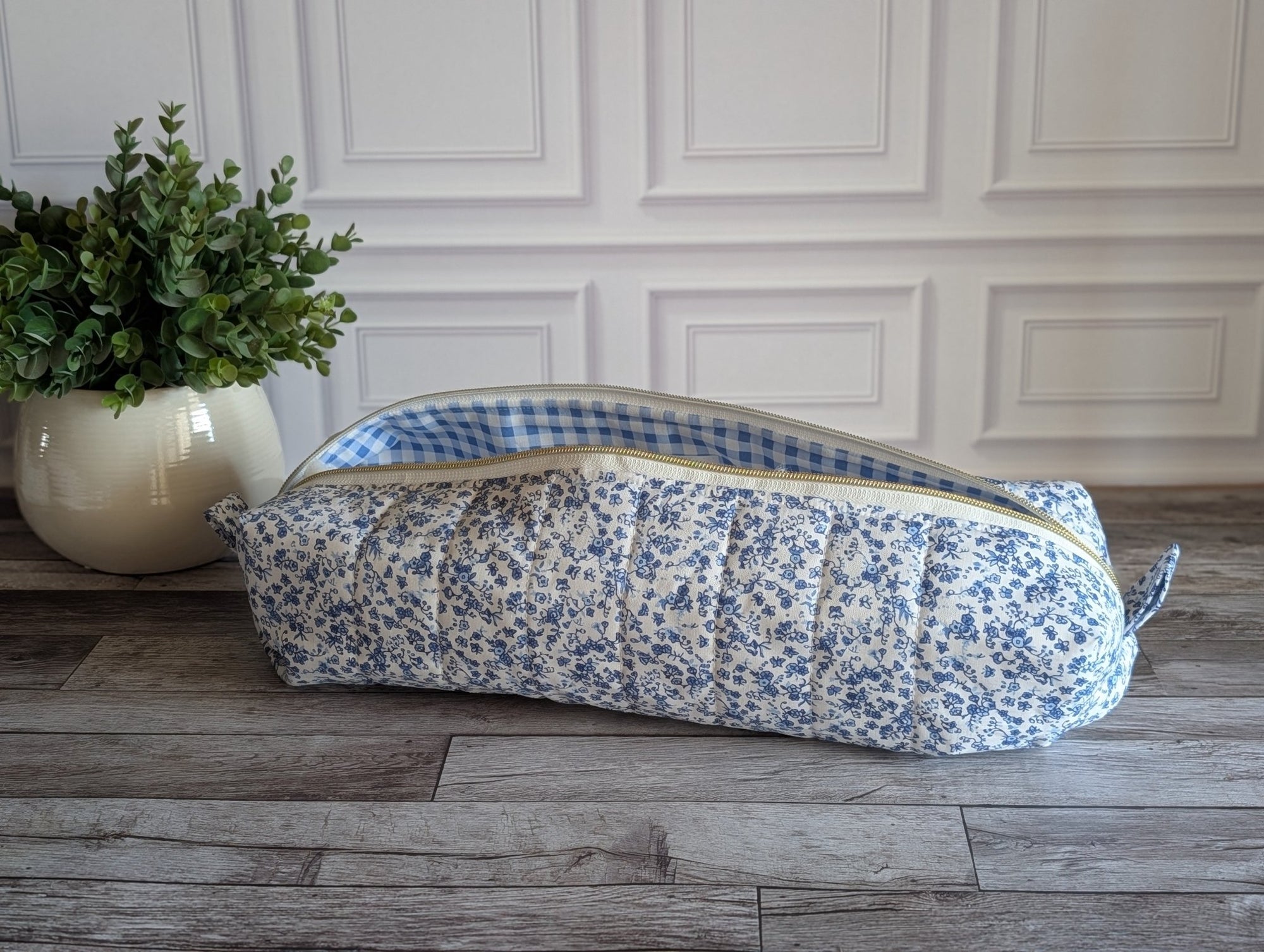 A quilted fabric zippered pouch with a blue and white floral print exterior and a blue-and-white gingham lining, resting on a gray wood floor next to a potted green plant against a white paneled wall.