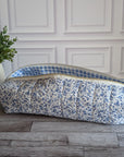 A quilted fabric zippered pouch with a blue and white floral print exterior and a blue-and-white gingham lining, resting on a gray wood floor next to a potted green plant against a white paneled wall.