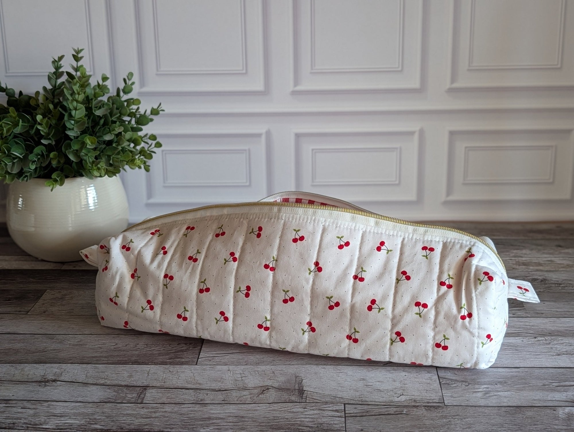 A quilted white cosmetic bag with a red cherry print, resting on a gray wooden floor in front of a white paneled wall next to a potted green plant.