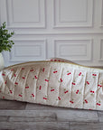 A quilted white cosmetic bag with a red cherry print, resting on a gray wooden floor in front of a white paneled wall next to a potted green plant.