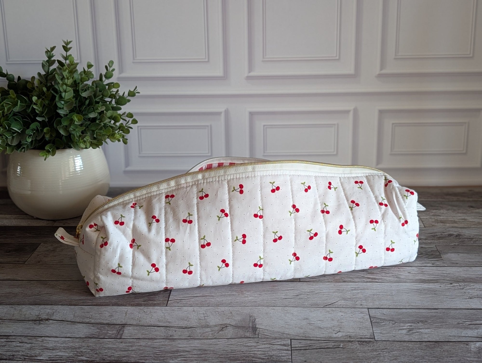A quilted white fabric bag with a red cherry print pattern, resting on a gray wooden floor in front of a white paneled wall with a potted green plant on the left.
