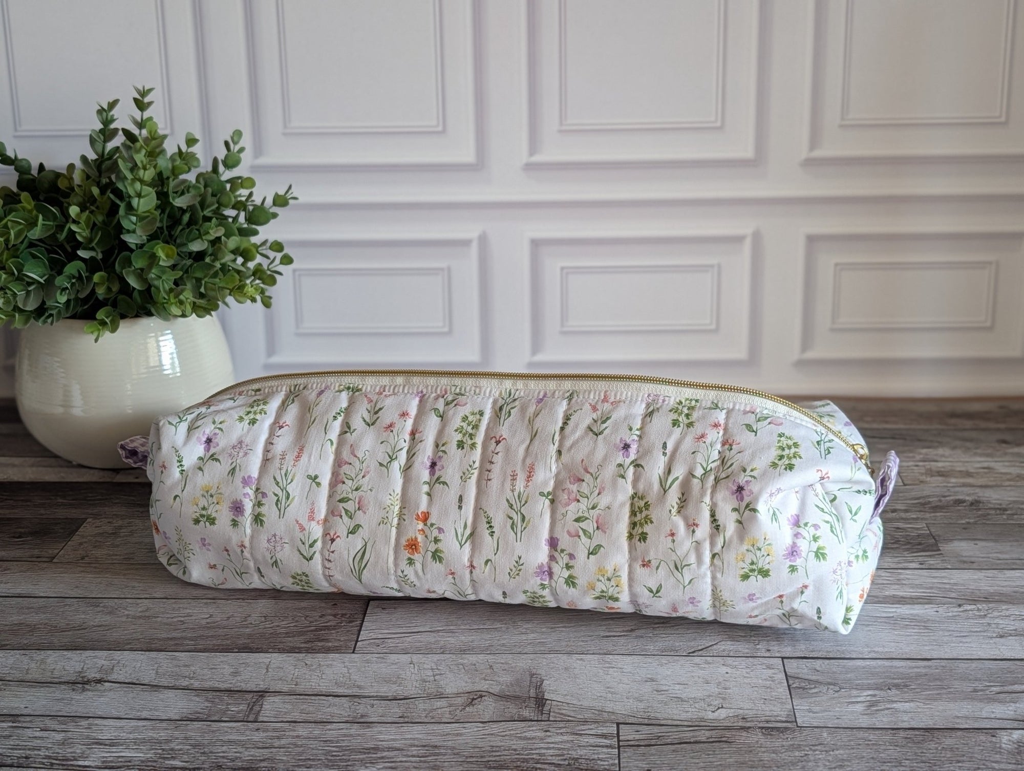 A quilted cylindrical cosmetic pouch with a delicate pink and green floral print, resting on a gray wood floor next to a potted green plant against a white paneled wall.
