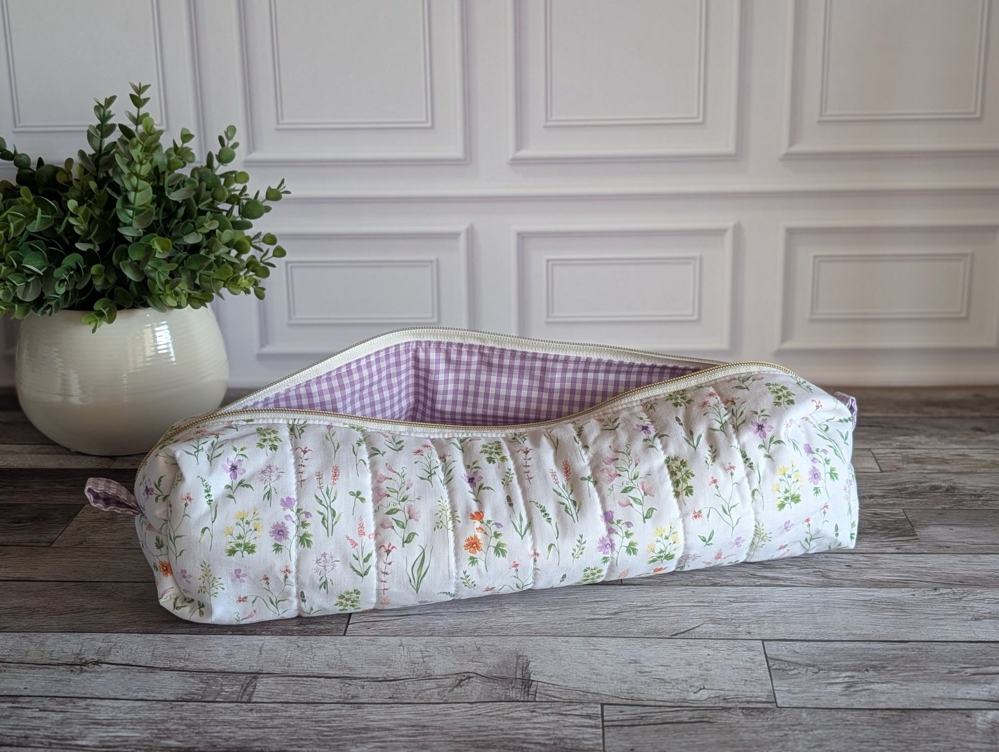 A quilted fabric pouch with a white floral print and a purple-and-white gingham lining, placed on a gray wood floor next to a potted green plant against a white paneled wall.