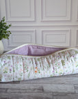 A quilted fabric pouch with a white floral print and a purple-and-white gingham lining, placed on a gray wood floor next to a potted green plant against a white paneled wall.
