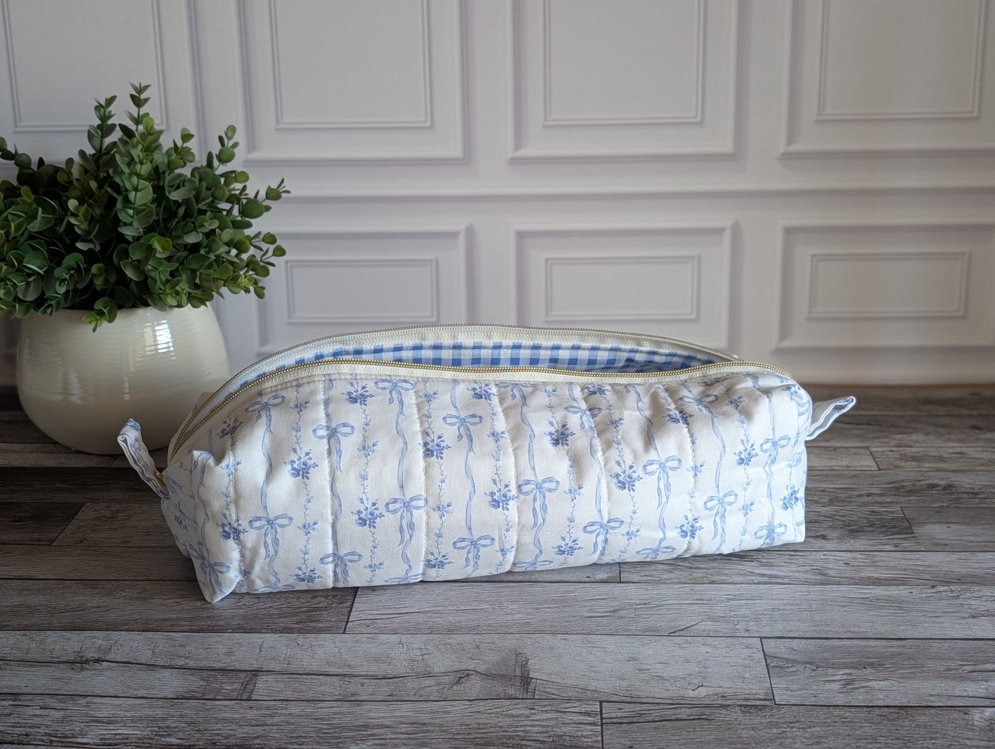 A quilted zippered pouch with a blue and white floral print and a blue gingham lining, resting on a gray wood floor next to a potted green plant.