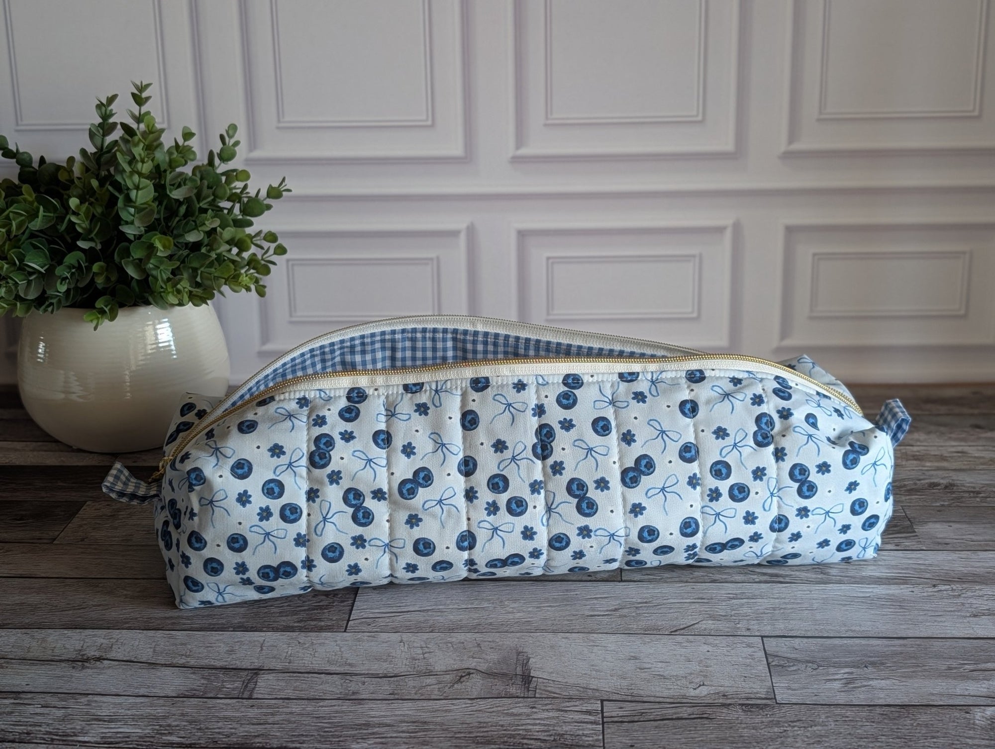 A quilted fabric zippered pouch with a blue and white floral print and a blue-and-white gingham lining, resting on a gray wood floor next to a potted green plant.