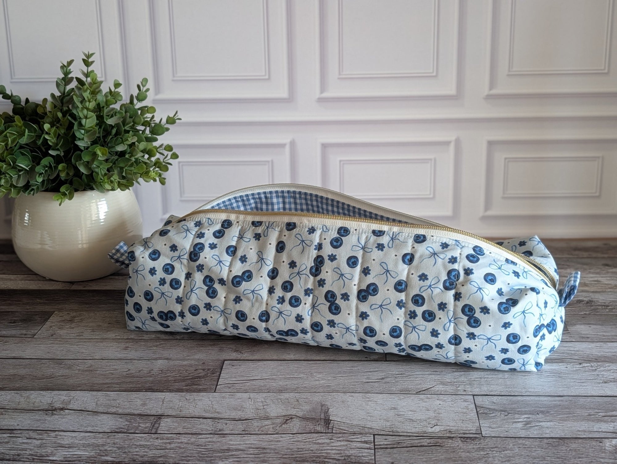 A quilted fabric zippered pouch with a blue and white floral print and a gingham lining, resting on a gray wood floor next to a potted green plant.