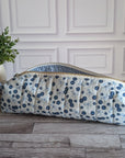 A quilted fabric zippered pouch with a blue and white floral print and a gingham lining, resting on a gray wood floor next to a potted green plant.