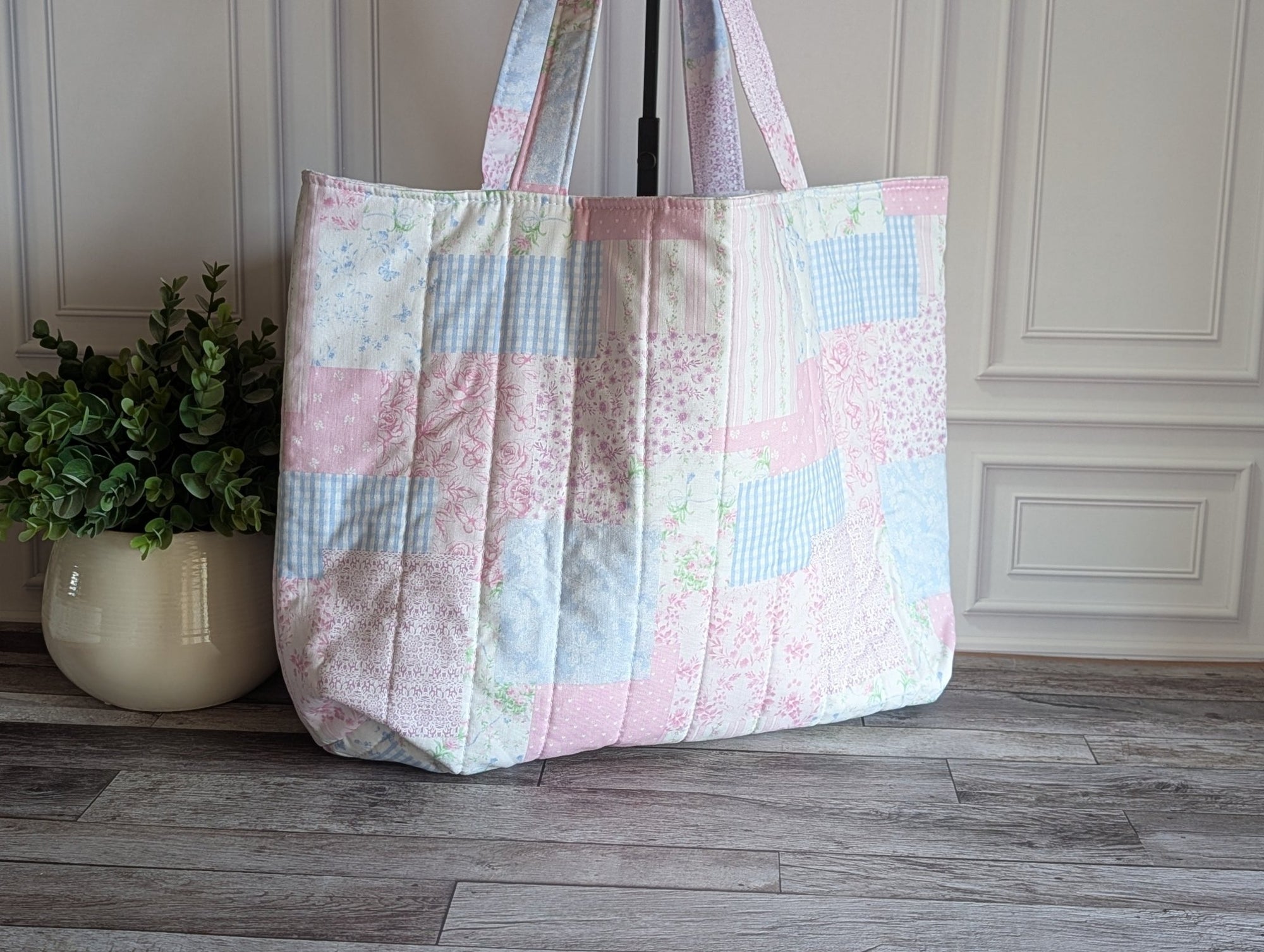 A quilted patchwork tote bag in pastel pink, blue, and white fabrics, standing on a gray wood floor next to a potted green plant.