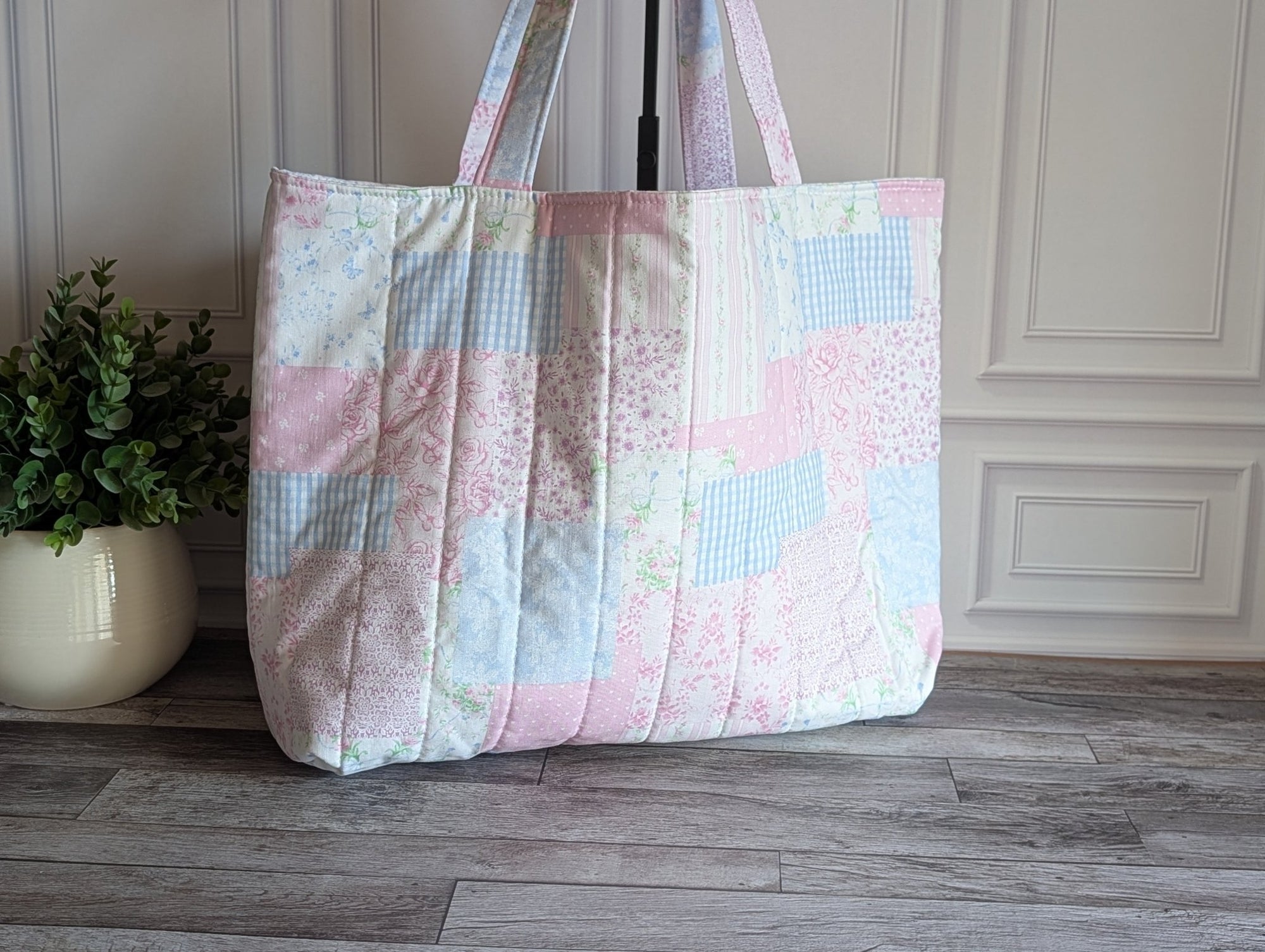 A large quilted patchwork tote bag in pastel pink, blue, and white fabrics, standing on a gray wood floor against a white paneled wall with a potted plant on the left.