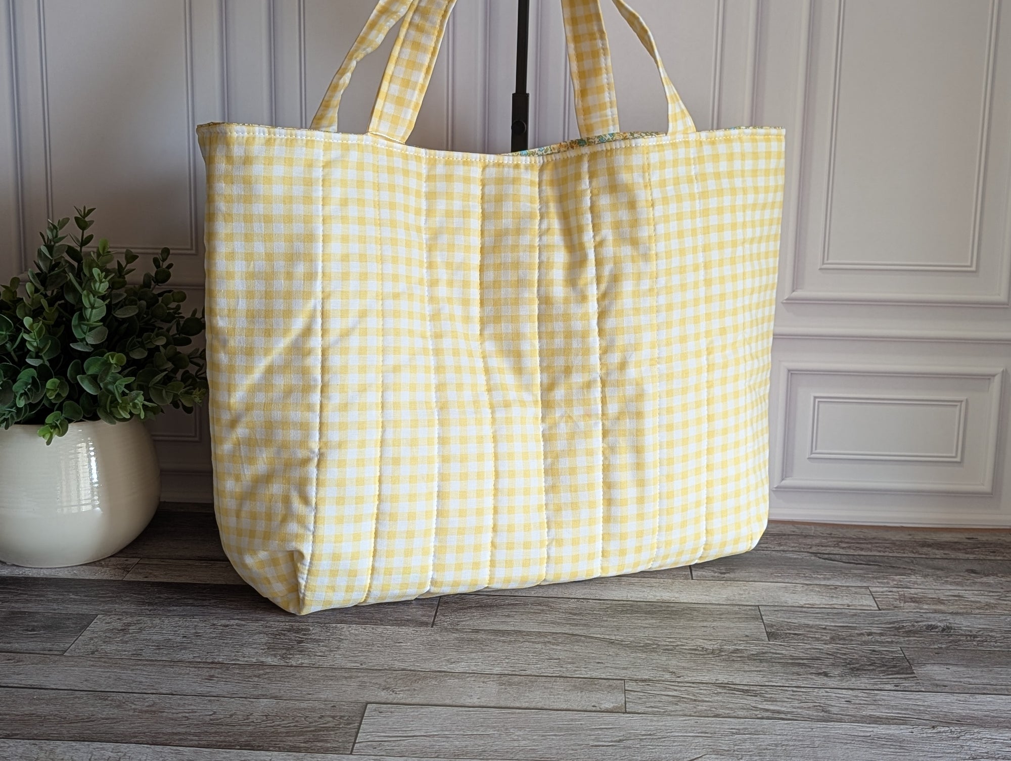 A large quilted tote bag with a yellow and white gingham check pattern, standing on a gray wood floor next to a potted green plant.