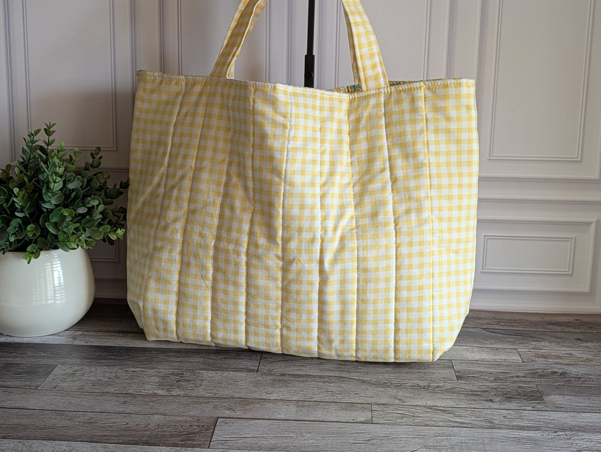 A large quilted tote bag in a yellow and white gingham check pattern, standing on a gray wood floor against a white paneled wall with a potted green plant on the left.