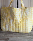A large quilted tote bag in a yellow and white gingham check pattern, standing on a gray wood floor against a white paneled wall with a potted green plant on the left.