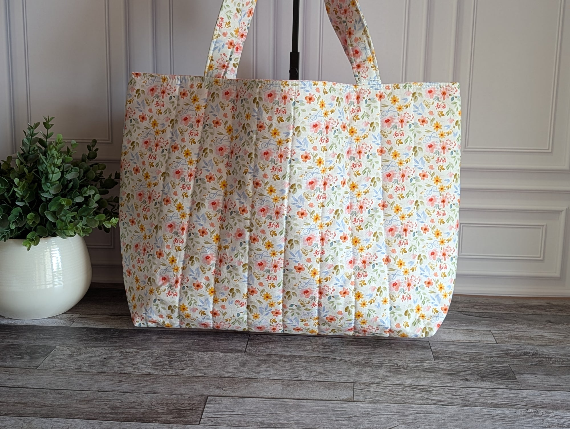 A quilted fabric tote bag with a small multicolor floral print, standing on a gray wood floor next to a potted green plant.