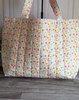 A quilted fabric tote bag with a small multicolor floral print, standing on a gray wood floor next to a potted green plant.