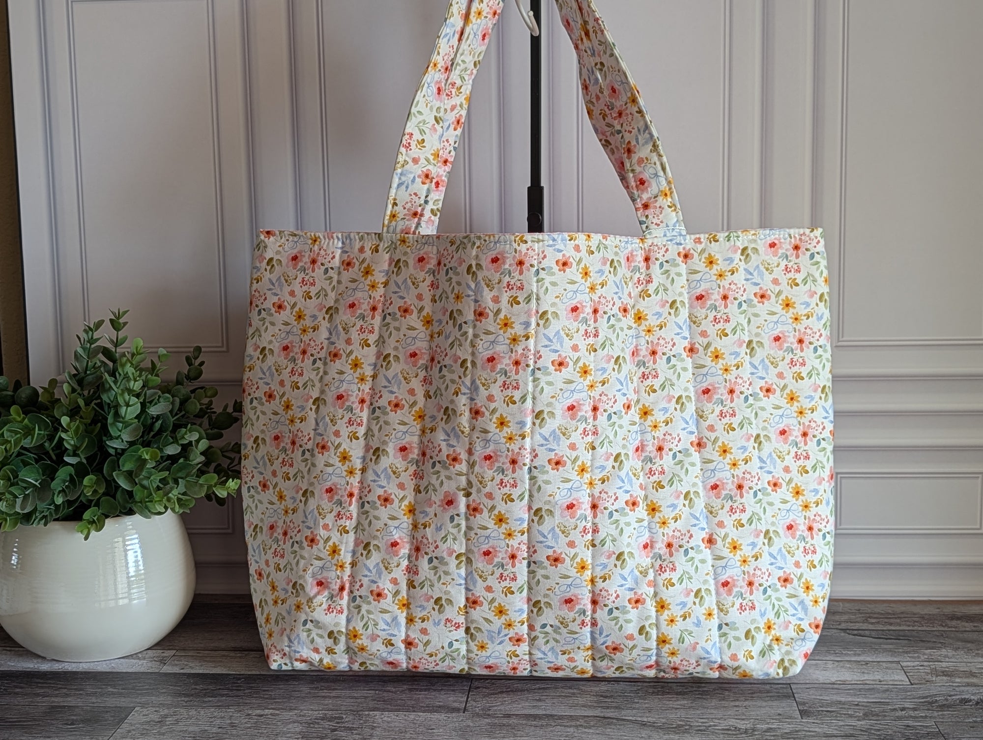 A large quilted tote bag with a small multicolor floral print on a cream background, standing on a gray wood floor next to a potted green plant.
