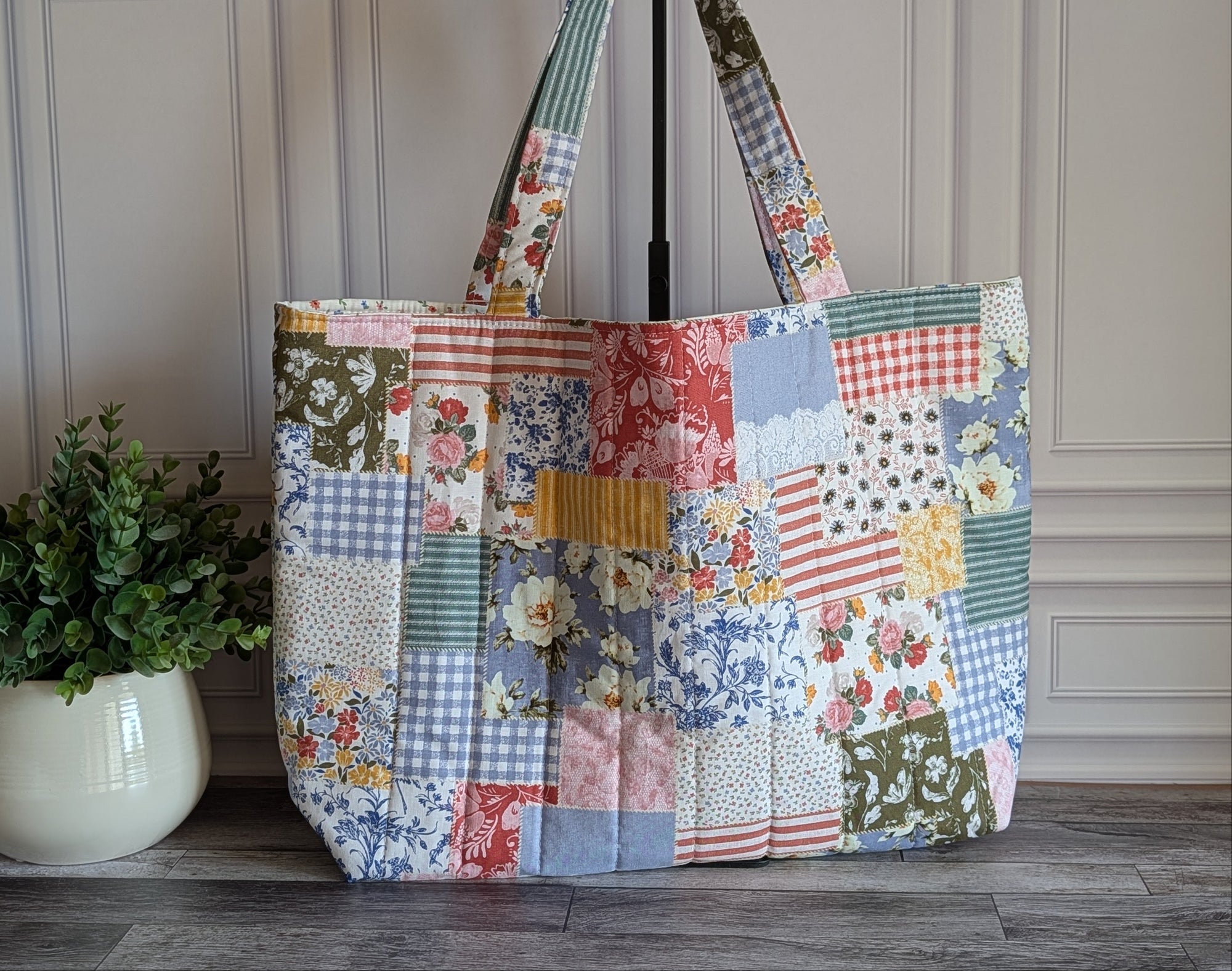 A large patchwork tote bag made from multiple vintage-style fabric panels in floral, gingham, and solid prints, displayed on a wooden floor next to a potted green plant.