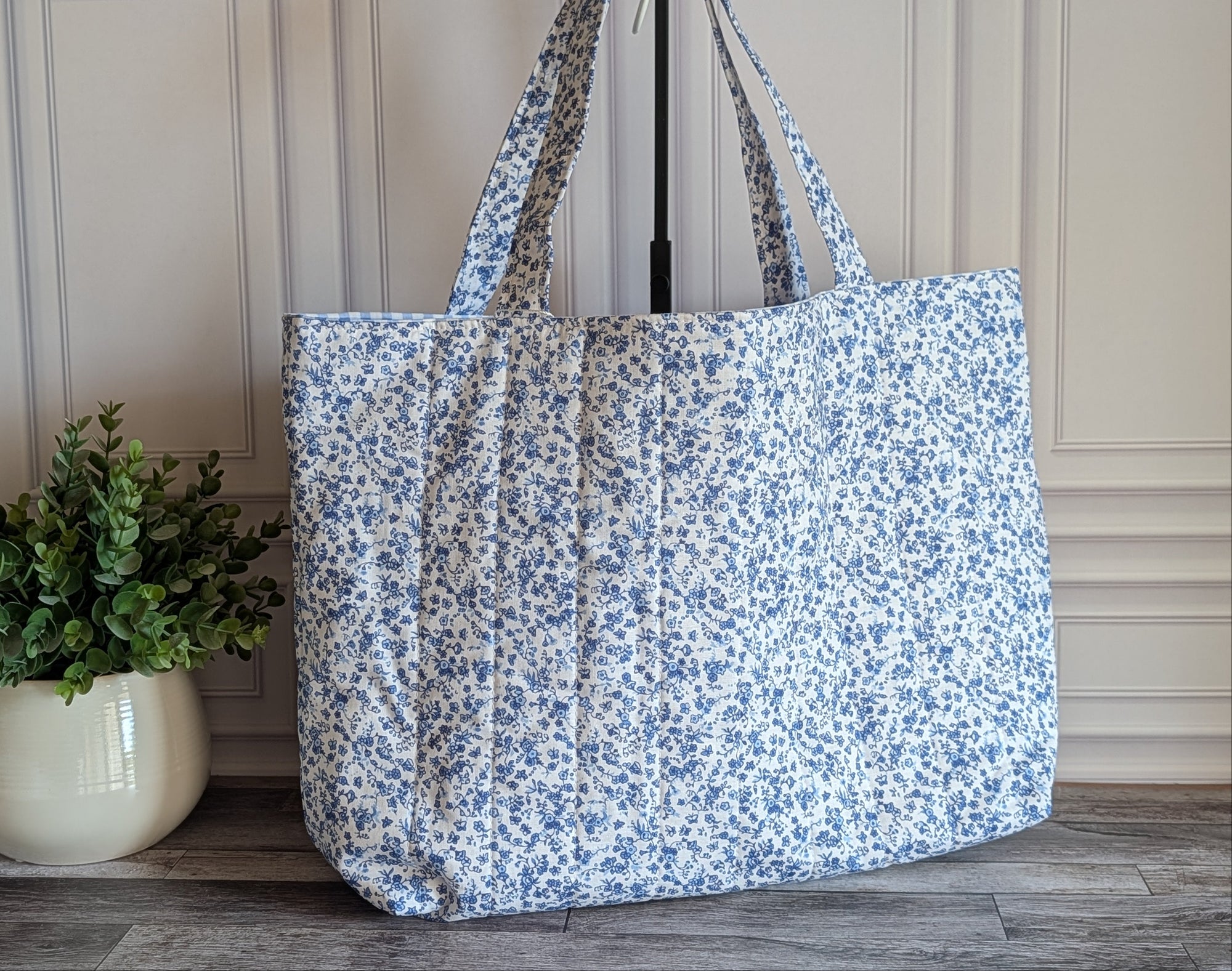 A large fabric tote bag with a blue and white floral print, standing on a gray wood floor next to a potted green plant against a paneled cream wall.