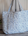 A large fabric tote bag with a blue and white floral print, standing on a gray wood floor next to a potted green plant against a paneled cream wall.