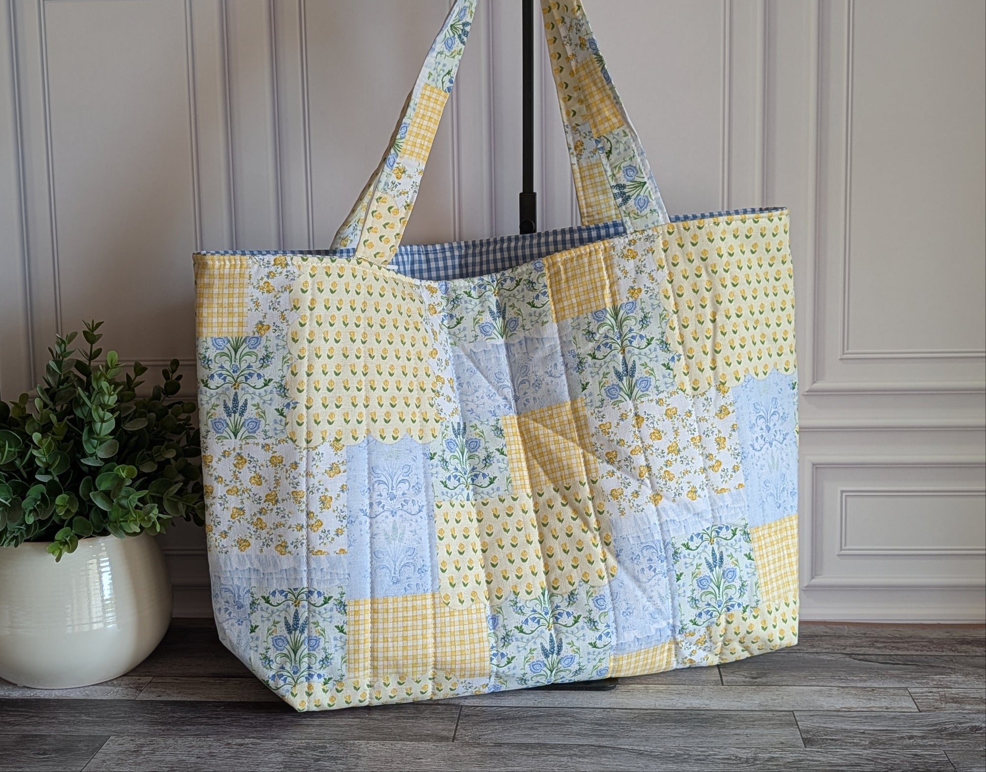 A large quilted patchwork tote bag with yellow, blue, and white fabrics, standing on a gray wood floor next to a potted green plant.